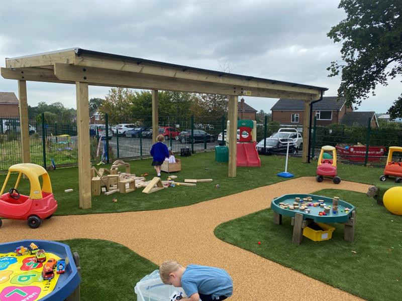 the timber canopy stands to the left in this picture as children play underneath it, you can see the green artificial grass and beige wetpour surfacing and the tuff spot table to the side as well as lots of toys
