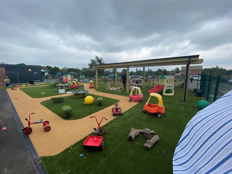 a photo showing various childrens toys such as ride-along cars and scooters lying across the beige wetpour roadway marked around sections of artificial grass
