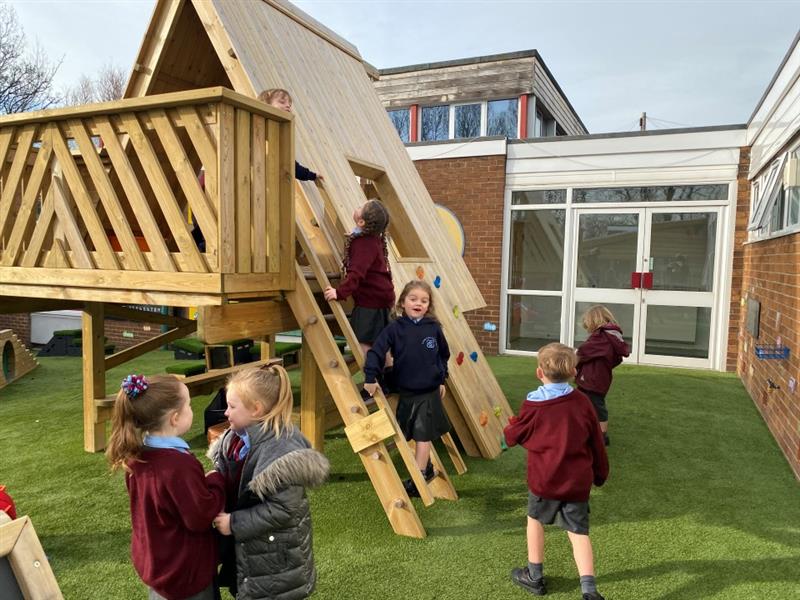 children in buguny school uniform and winter coats stand on the green artificial grass surfacing and look up at the timber learning den