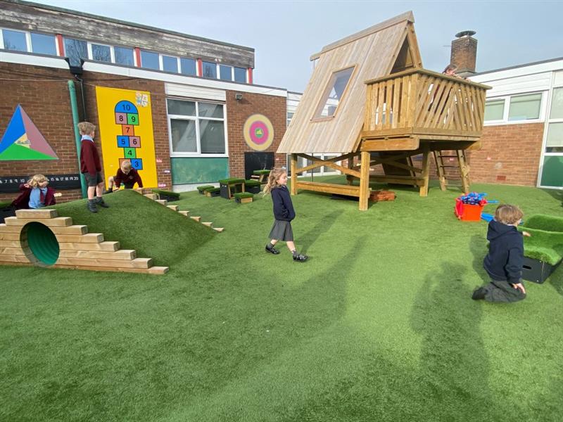 the artificial grass playturf ont he ground with the tree top learning den in the back and the playground mound to the side
