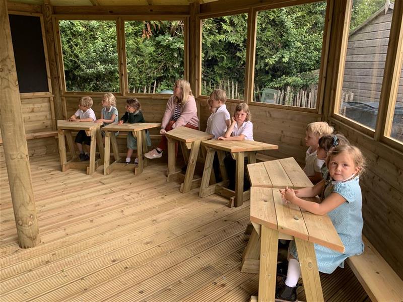 Inside the gazebo at hollycombe primary school, 9 children looking at the chalk board and one teacher accompanying them.