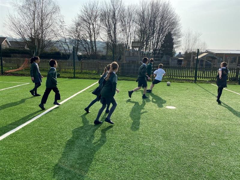 children in black and white school uniform kick the football around on the green artificial grass on the muga pitch
