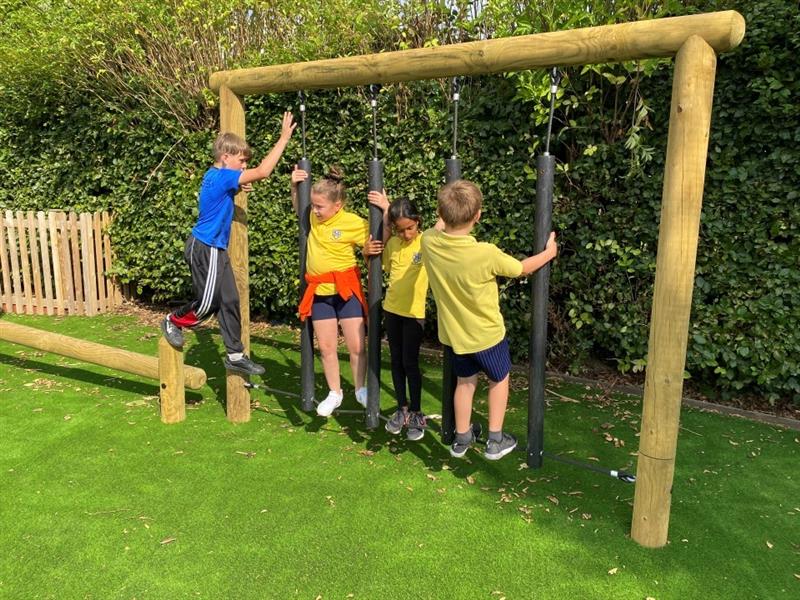 four children in brightly coloured clothing cross the rubbing stepping traverse as part of their key stage two trim trail, there is luscious green artificial grass 