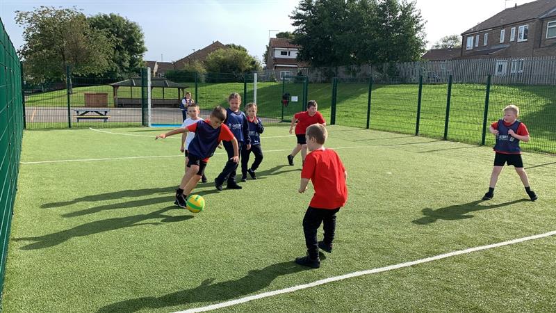 children in pe kit play on the green artificial grass surfacing within the muga with a football