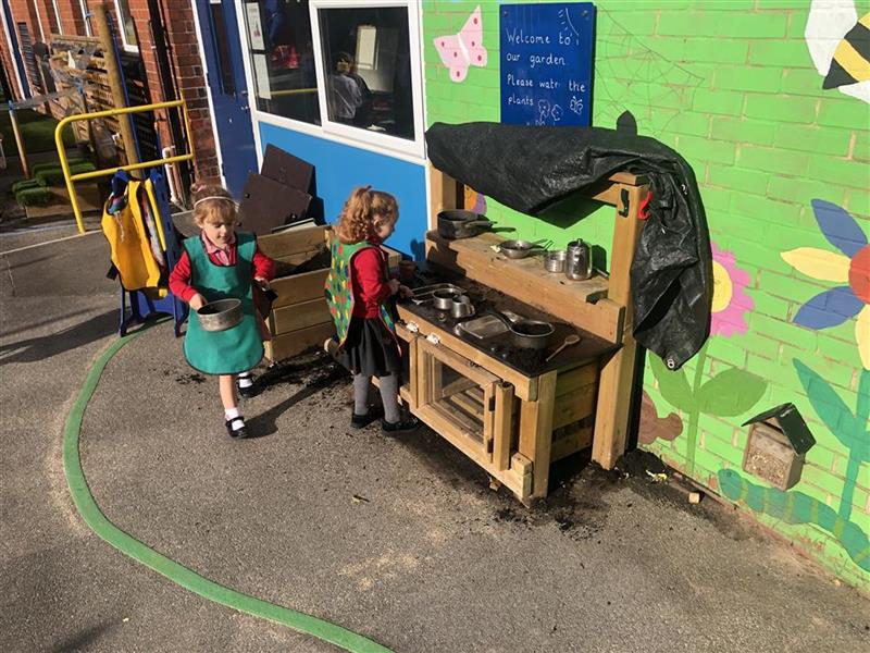 children in green aprons and red school uniform lay with a timber mud kitchen carrying pans of mud 