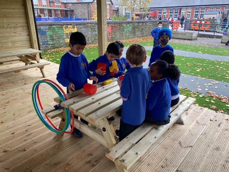 a group of little boys in royal blue school jumpers and black trousers gather in the outdoor classroom around a timber picnic table and play with coloured blocks, there are colourful hula hoops to the side also but they are not being played with