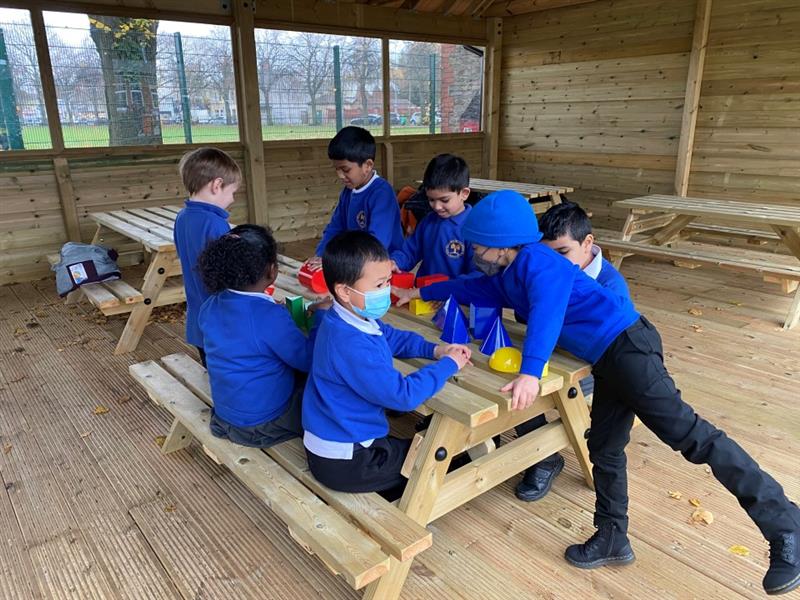 children in blue and black school uniform gather around a timber picnic table within the outdoor classroom playing with colourful toys on the table