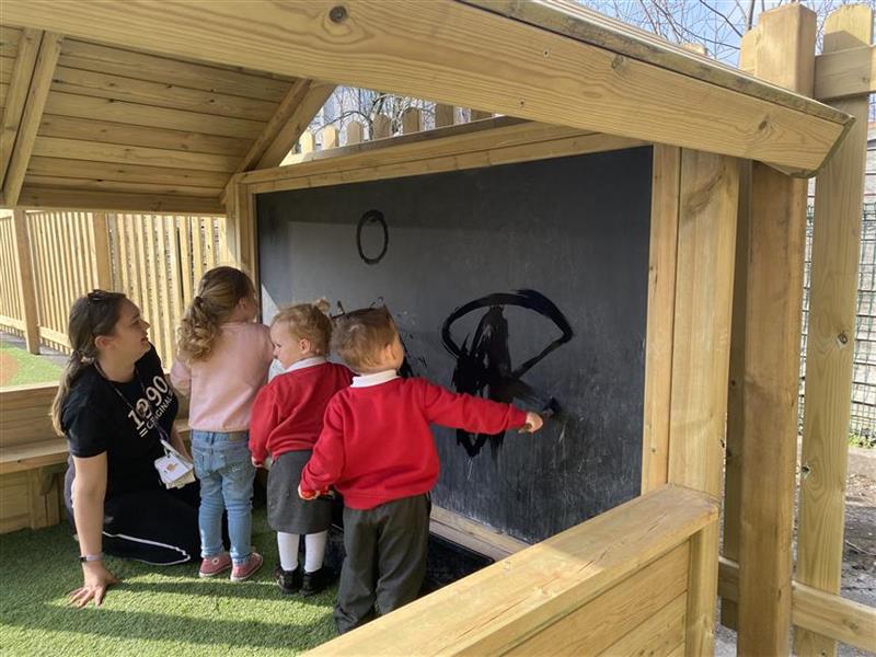 3 children and one teacher are sat in the play house, drawing on the giant chalk board