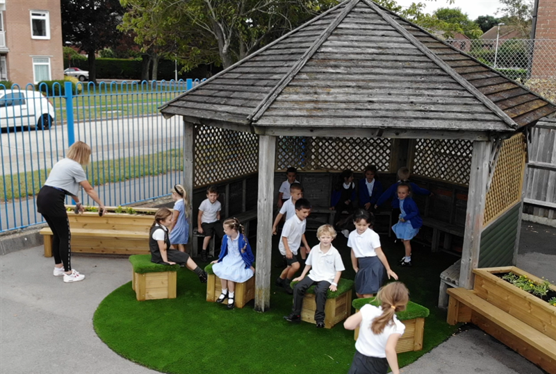 Artificial grass surrounding the old canopy at Thomas A Beckett Primary School. Children are sat on top of the grass top seats.