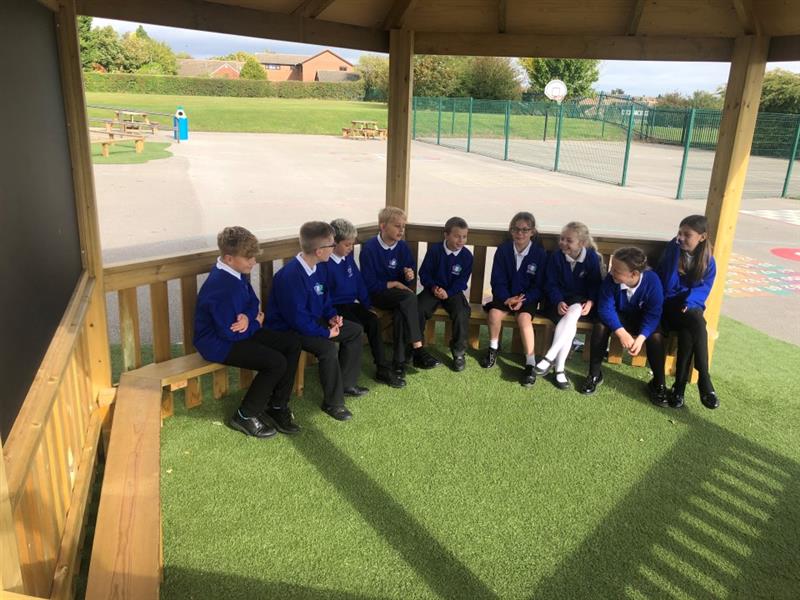 Children sit patiently in a hexagonal gazebo wearing a blue, white and black school uniform chatting to one another, there is a giant chalkboard in the corner of the hexagonal gazebo