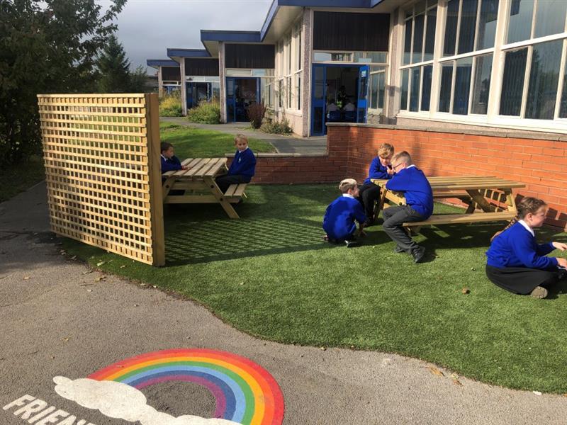 children gather around the picnic tables on the artificial grass surfacing with the timber trellis fences and a friendship rainbow playground marking 