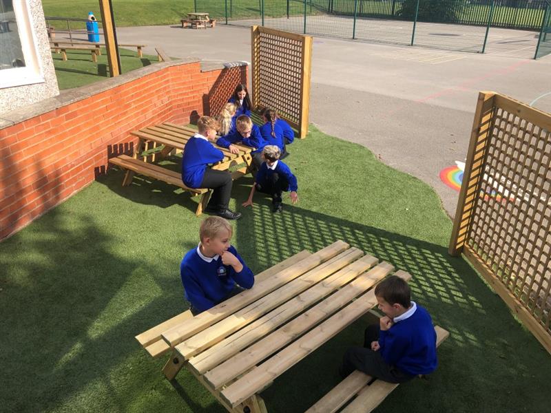 children gather around sitting and chatting over a picnic table with green artificial grass surfacing below, this is a dedicated quiet area