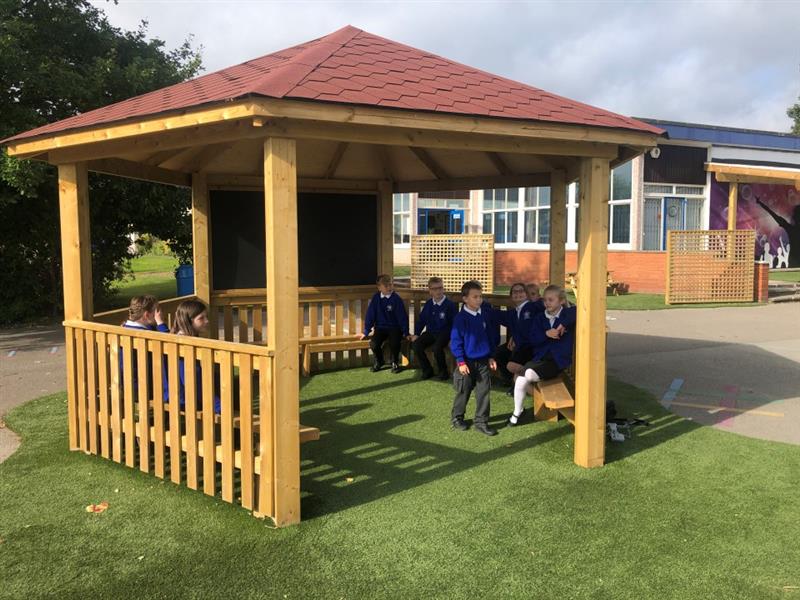 a view of the outside of the hexagonal gazebo on artificial grass playturf with children in blue school uniform sat on the benches inside, a little boy walks around instead of sitting down