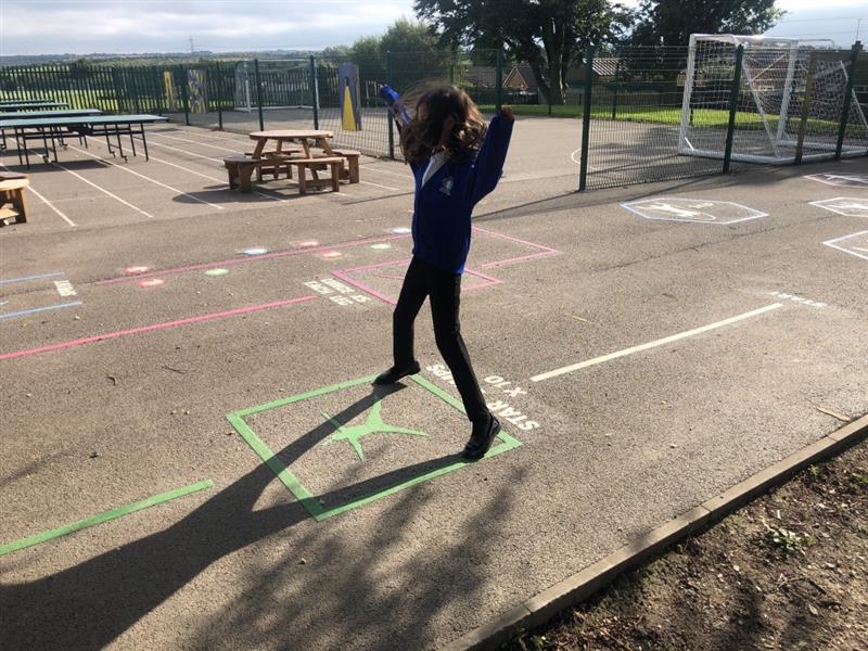 a child in blue, white and black school uniform runs across the grey tarmac playground on the green playground markings that instruct them to do a star jump