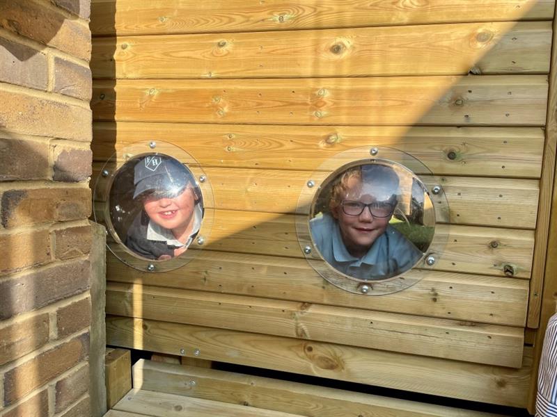 children peer through the window of the lookout cabin towards the camera, one is wearing a blue/grey cap and the other is wearing glasses, they are both wearing light blue school uniform