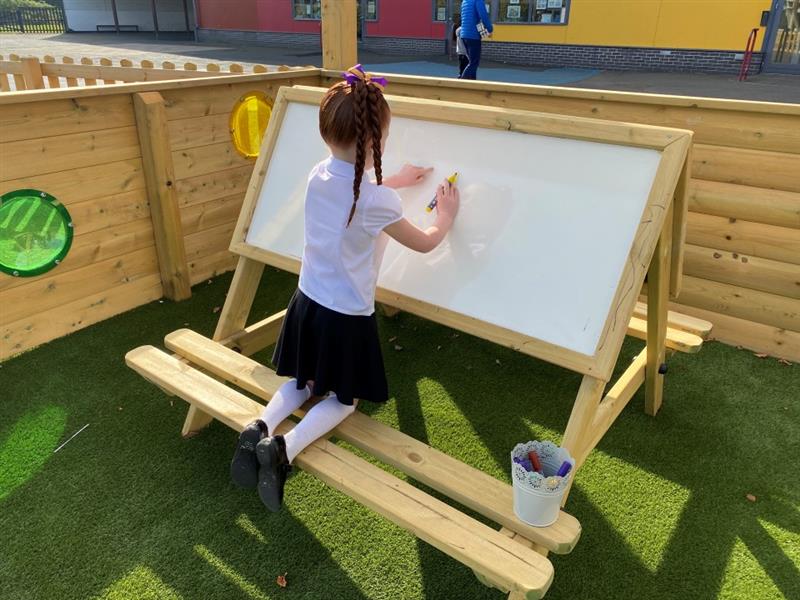a child sits at an easel table and draws on the whiteboard side underneath the timber canopy