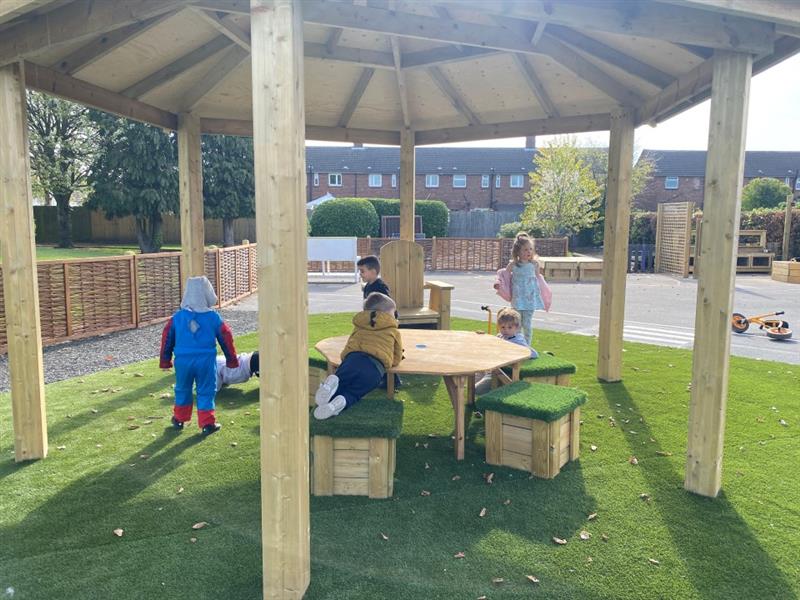 children sit underneath the hexagonal gazebo sitting on the artificial grass topped seats and the storytelling chair 