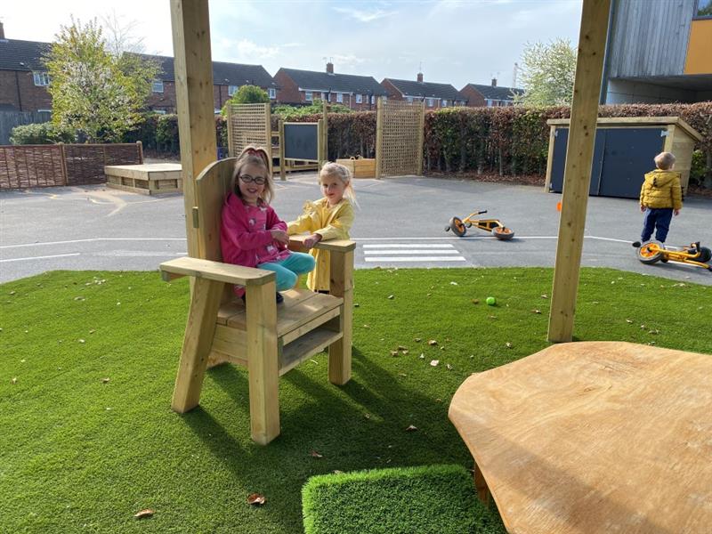 a child sits in the story-telling chair and smiles at the camera whilst her friend in a yellow coat stands behind her