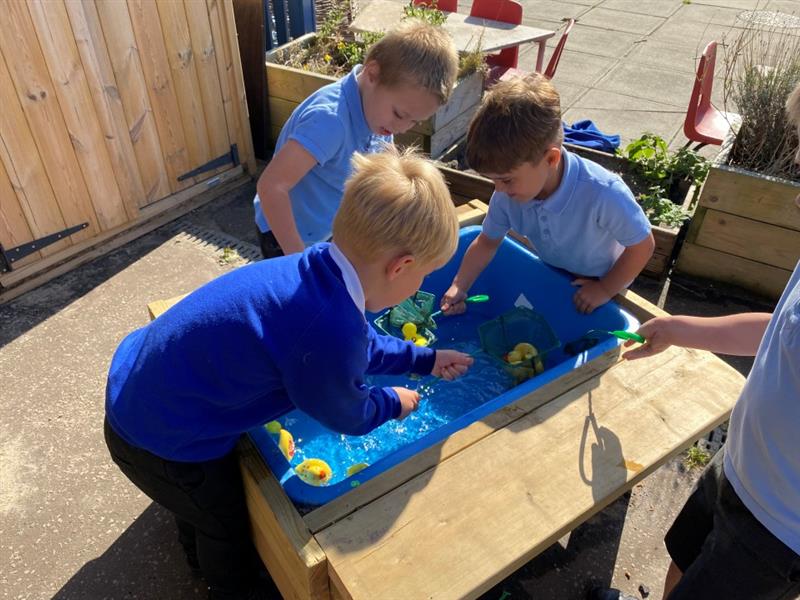 three little boys in blue and black school uniform are playing with water in a timber and blue plastic water tray