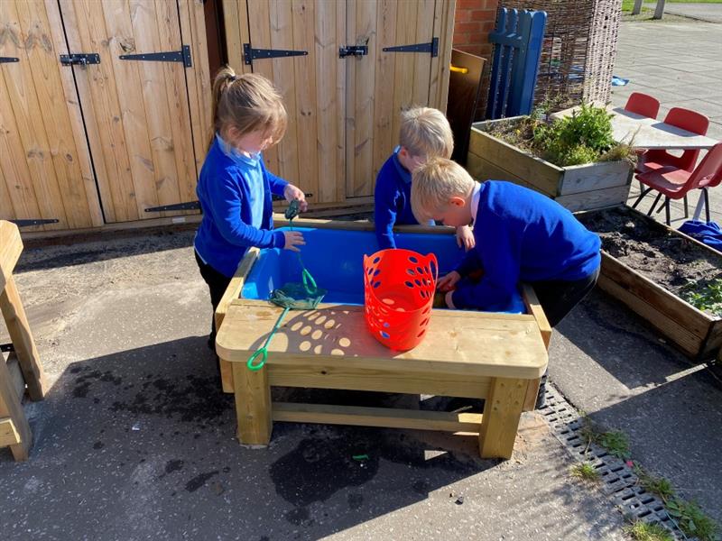 a little girl and two little boys in blue nd black school uniform gather around a water table with lid ad play with the water and toys in the water