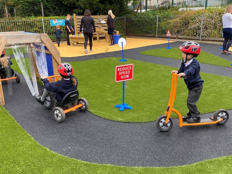 two children in school uniform drive around the black roadway track on orange scooters and trikes and through a wooden play car wash