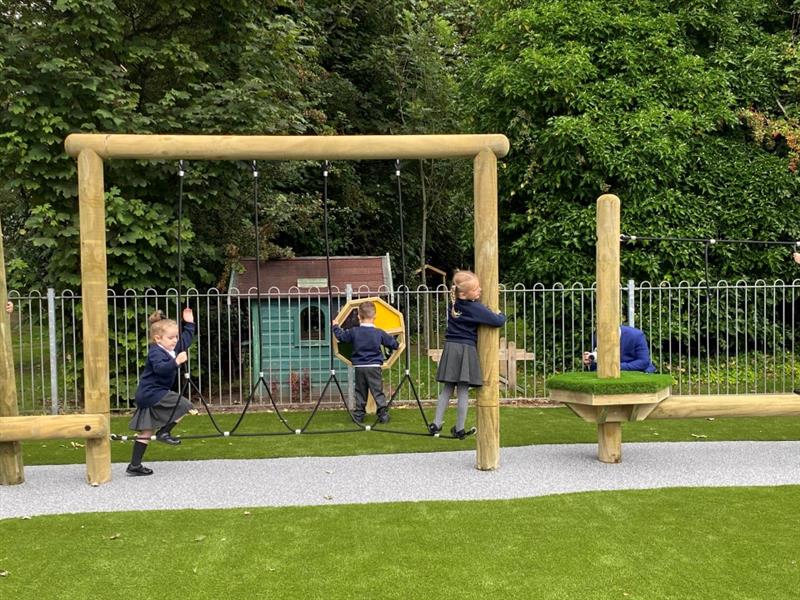 four children scale the wistman's forest trail, two children cross the net rope traverse, one child leans across from the balance beam to the artificial grass topped platforms and one child plays with the sensory wheel behind the trim trail