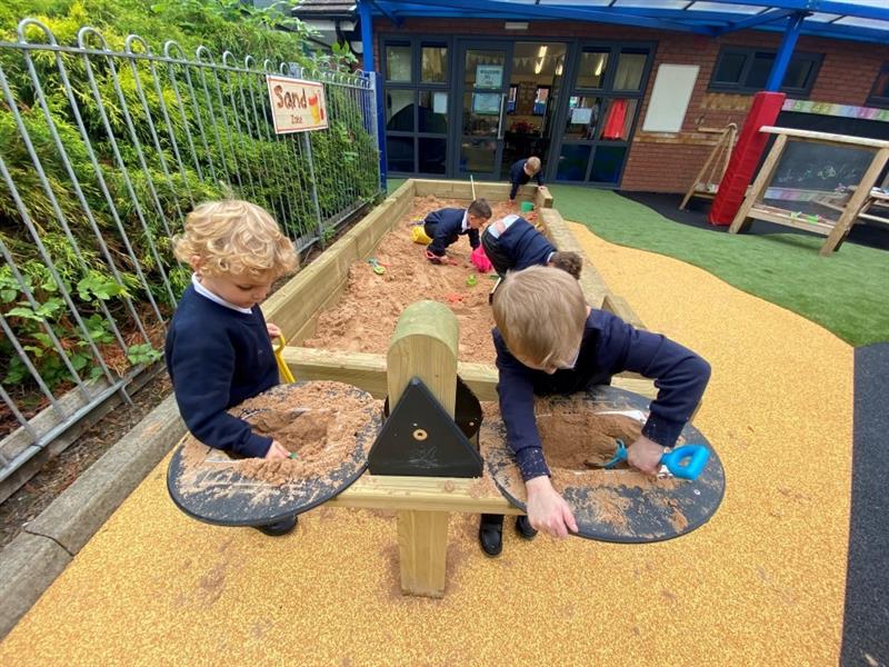 two little boys in blue school uniform have their heads down filling the black weighing scales with sand using spades and their hands