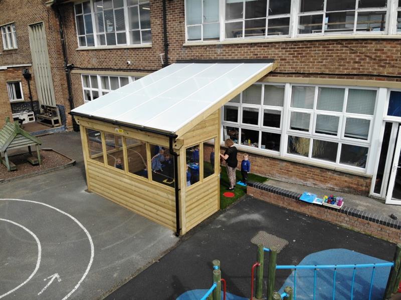 Children sit in the canopy on the artificial grass under the shelter whilst being guided by their teacher
