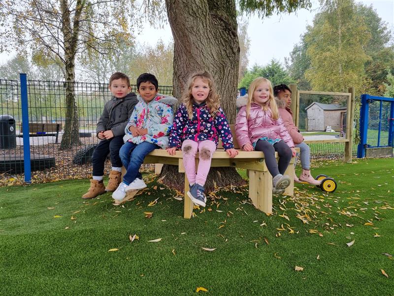 five children in winter coats sit on the octagonal tree bench looking at the camera with the weeping willow tree behind them