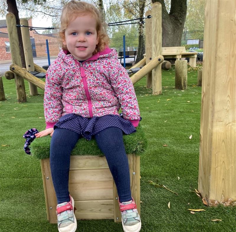 a little girl in a pink winter coat sits on the artificial grass topped seat and smiles at the camera