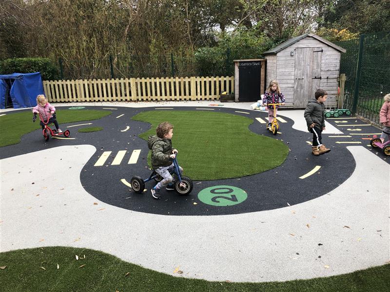 four nursery children in winter coats play on scooters on the wetpour roadway