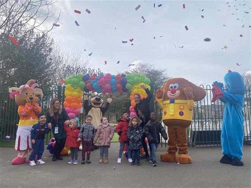 4 mascots, 7 children and 2 teachers are stood in front of balloons with a confetti cannon going off behind them.