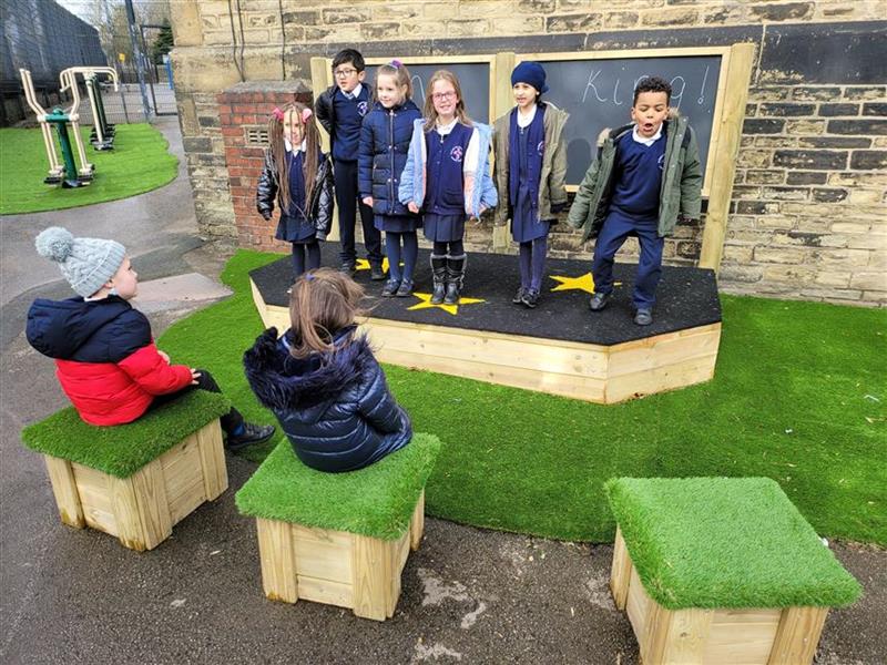 6 children playing on a performance stage whilst 2 pupils are watching them sat down.