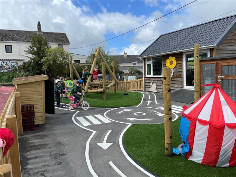 a whole view of the playground with a grey roadway, artificial grass, timber den making posts and a climber