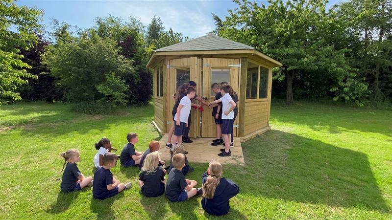 children open the doors to the new outdoor classroom for their peers who are sat on the grass in front of the outdoor classroom