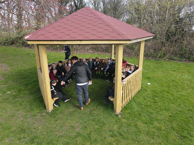 the hexagonal gazebo sits on the green grass of the field whilst the children sit on the benches