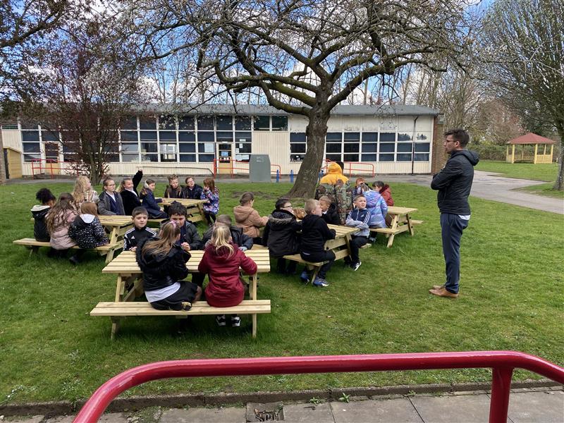 children sit on the timber picnic tables and listen to their teacher teaching them a lesson outdoors