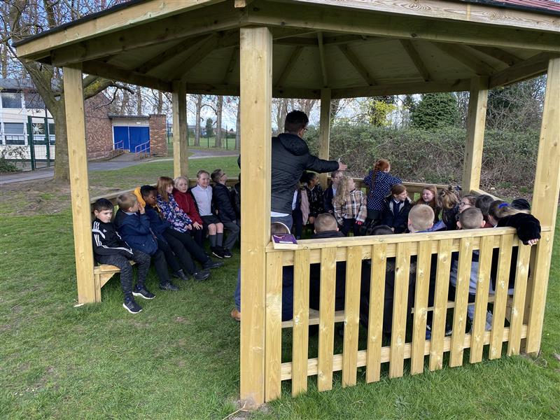 children sit inside the gazebo on the benches and listen to a teacher leading a lesson