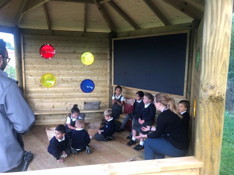 children within a sensory gazebo on the floor and on timber benches,, they wear black and white school uniform and there is a giant chalkboard against one of the walls, there are also colourful sensory bubble windows in red, yellow and blue