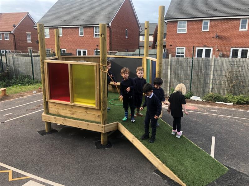 five pupils in black and white school uniform stand on an artificial grass surfaced ramp and look at the camera and the floor respoectively as they wlak up to the sensory panels surrounding the little imagination station den in their playground