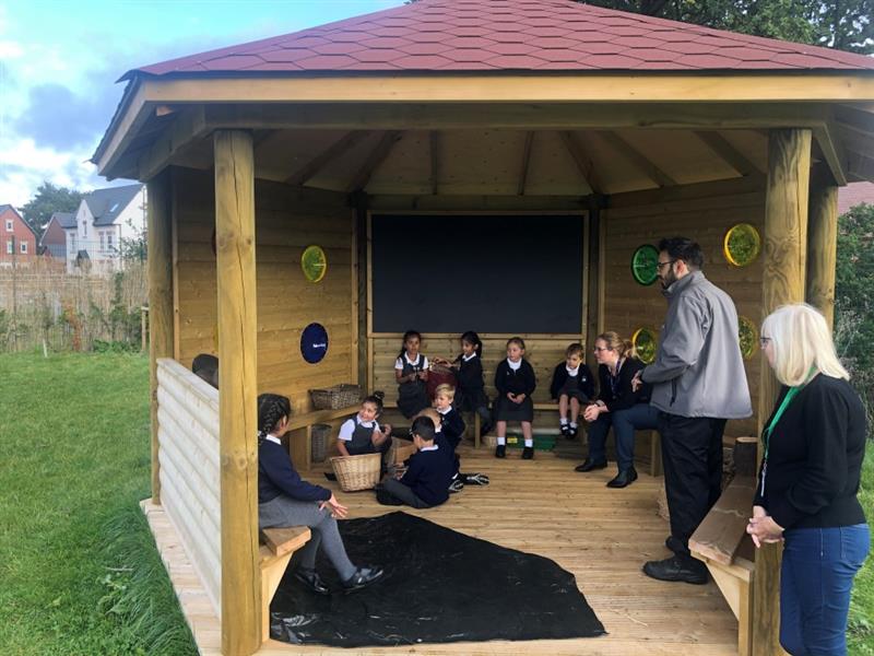 a teacher leads a class in the sensory gazebo as most children sit gathered around in a semi circle on benches and some sit on the floor, the teacher is wearing a big grey jumper and the children wear a black and white school uniform