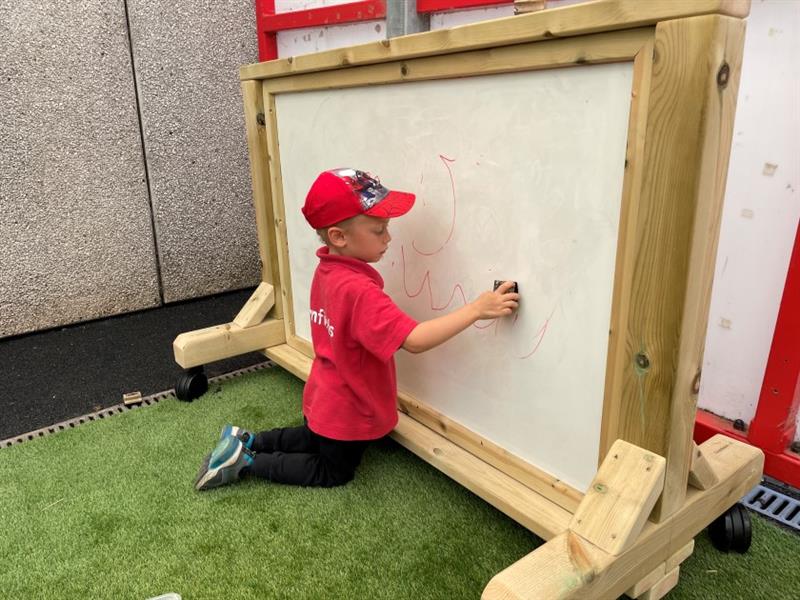 a child in a red and black uniform kneels on the grass and draws on the whiteboard 