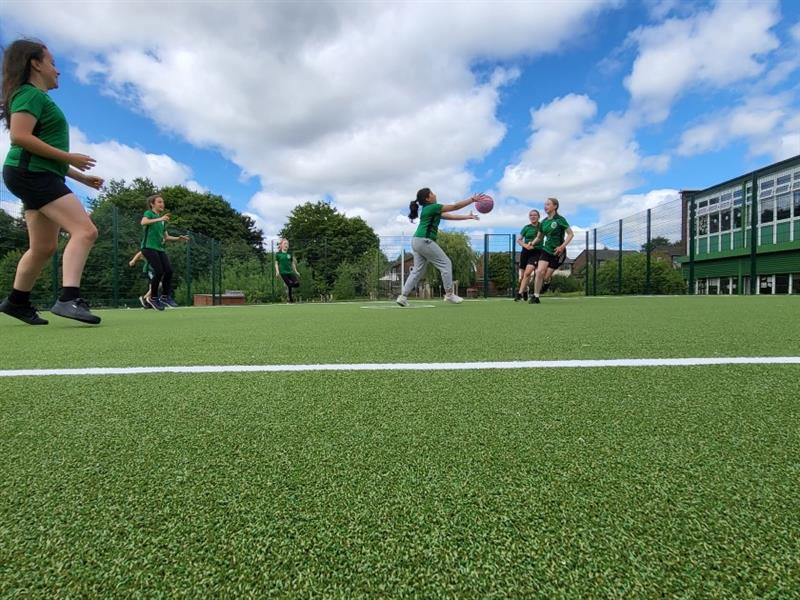 children play basketball together as part of a pe lesson on the green artificial grass muga