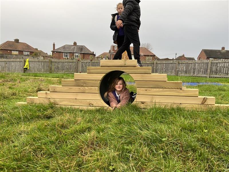 2 children climbing over the climb through tunnel hill in their playground setting whilst one girl is going through the tunnel