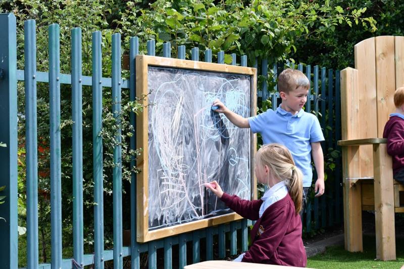 a little boy stands and draws on the chalkboard whilst a younger girl kneels on the floor drawing on the bottom of the board