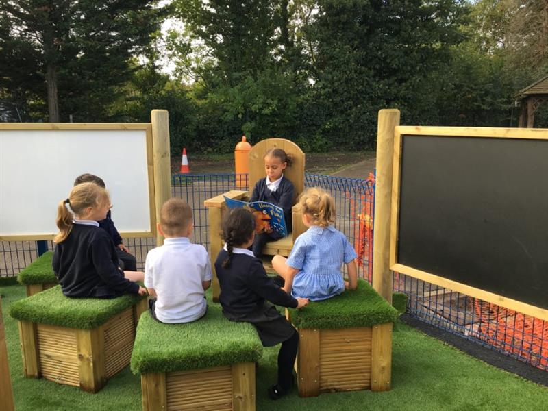 a child sitting on a storytelling chair reading out loud to 5 children sat on artificial grass topped seats