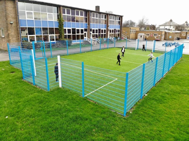 A 20m x 12m muga with blue sport fencing and artificial grass surfacing installed onto a school field with 5 children playing football
