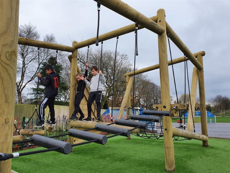 3 children playing on Pentagon Play's Puzzlewood Forest Circuit installed on the school playground with Artificial Grass underneath
