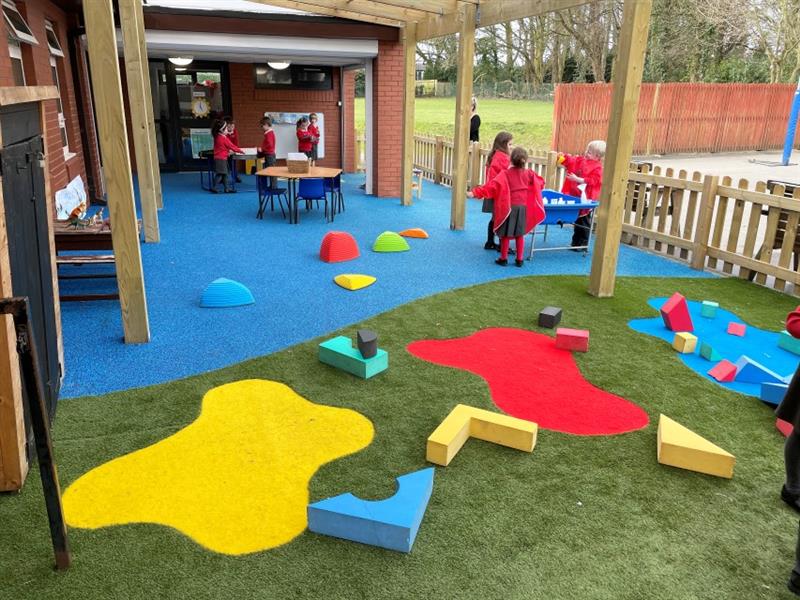 Eight children wearing red school jumpers are playing underneath the timber canopy on the blue wetpour surfacing which has been installed next to the school building whilst one teacher supervises. 