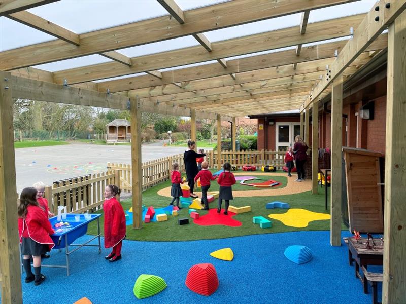3 children wearing red waterproof overalls stood at a water table that has been placed onto blue wetpour surfacing whilst 3 children speak to a teacher stood on the artificial grass and one child speaks to a teacher on the wetpour roadway. 